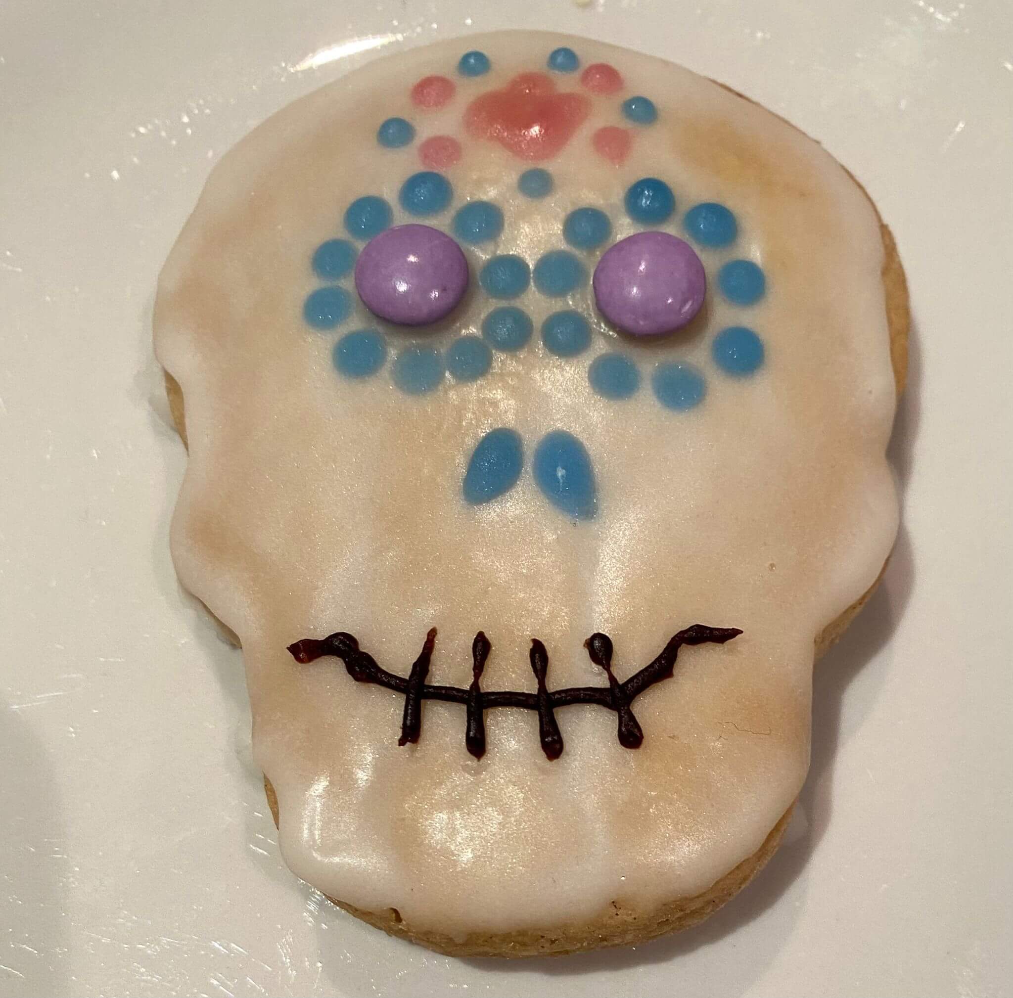White iced cookie in the shape of a skull. It is decorated with a black mouth, blue nostrils, purple eyes, a red and green flower on it's forehead and blue flowers around it's eyes.