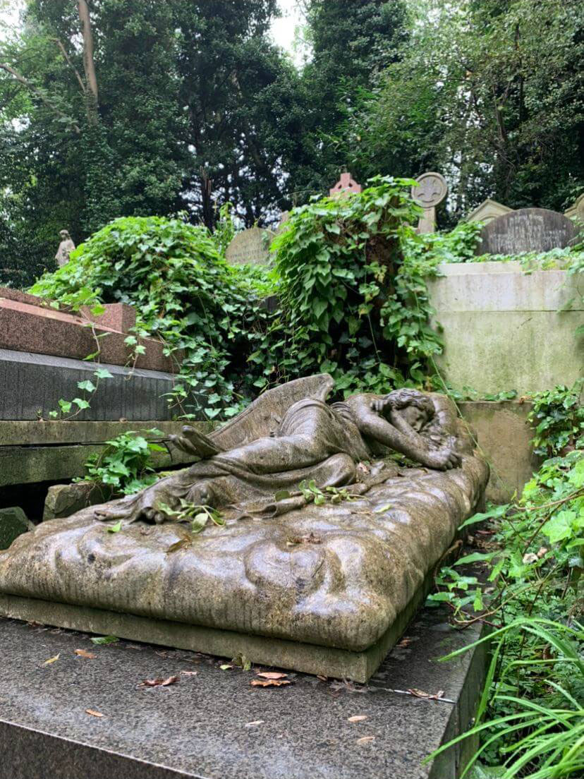 A statue of an angel lying down in a sleeping position is on top of a long rectangular grave. The grave is surrounded by other graves, with ivy growing on top of them. The graves are surrounded by tall trees.