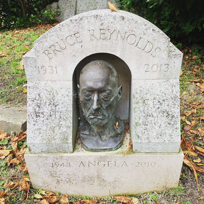 A bust of Bruce Reynolds is in the middle of this arched grave in a graveyard. The words above the bust read, 'BRUCE RENOYLDS 1931-2013\'. Below the bust reads, '1948 ANGELA 2010\'.