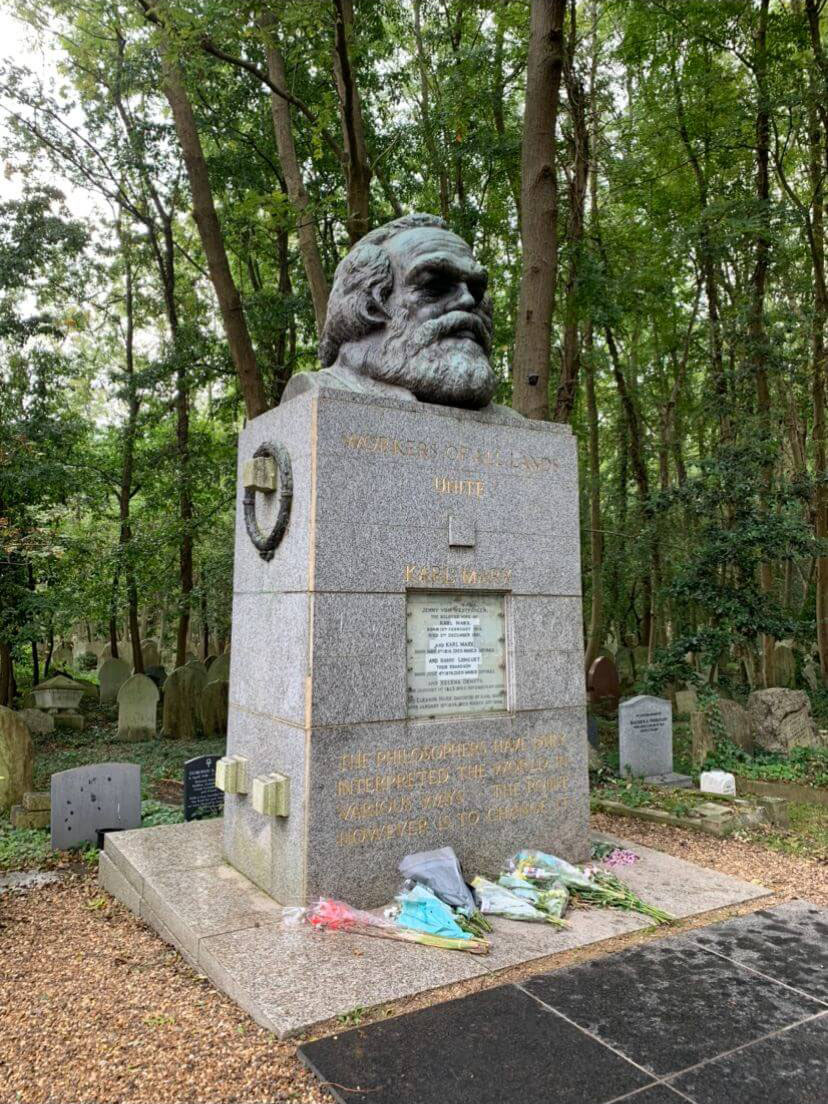 A bust of Karl Marx is on top of a cuboid shaped grave. There is a black stone path to the grave, and flowers in front of the grave. There are other smaller graves behind the Marx grave and are surrounded by trees.