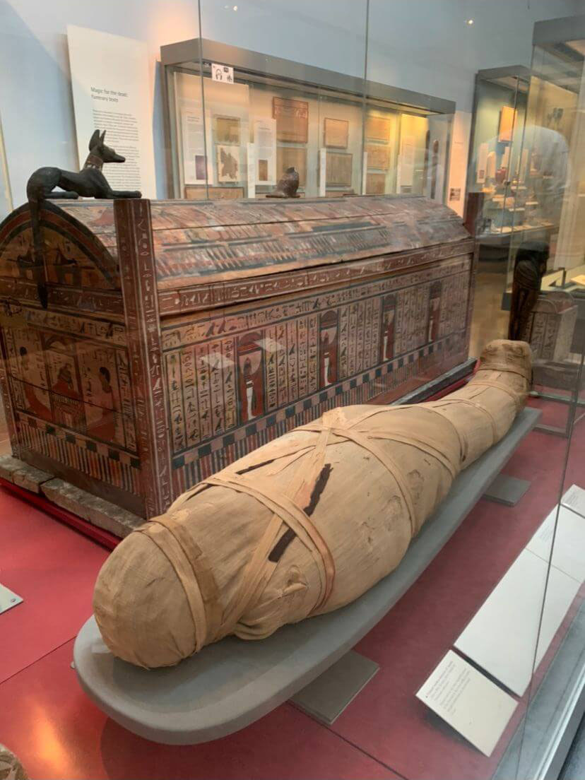 A mummy wrapped in various cloth is laying down on its\' back, with its\' feet furthest away from the camera. Behind the mummy is the sarcophagus/coffin made of wood. It is decorated in Ancient Egyptian hieroglyphics and paintings, with a statue of Anubis on the top.